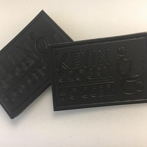 Puede incluir: Dos parches rectangulares negros con el texto en relieve "KEVIN GROGAN SOCCER". Los parches tienen esquinas redondeadas y un borde en relieve. El texto está en una fuente estilizada.