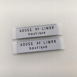 May include: Two rectangular woven labels with the text "HOUSE OF LIMAR boutique" in black letters. The labels are light gray with white edges, and are likely for clothing or accessories.