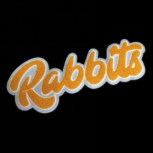 以下が含まれることがあります： 筆記体でRabbitsという文字がデザインされたシェニール織のワッペン。文字は鮮やかなゴールデンイエローで、厚みのある質感のパイル地となっており、無地の黒い背景に対してライトグレーの縁取りが施されています。