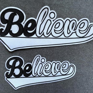 May include: Two white and black embroidered patches with the word "Believe" in a cursive font. The patches are shaped like a baseball team's logo.