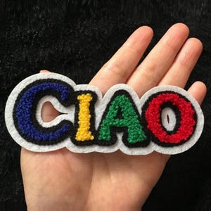 Puede incluir: Un parche bordado colorido con la palabra "CIAO" en una fuente de estilo universitario. Las letras son azules, amarillas, verdes y rojas, delineadas en negro, sobre un fondo blanco.