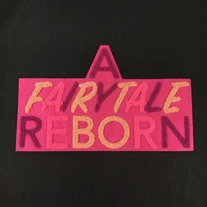 Op de afbeelding: Een rechthoekige roze wandhanger met de tekst A FAIRYTALE REBORN in verhoogd, getextureerd punch needle borduurwerk. De letters wisselen af tussen perzik, lichtroze en donkerpaarse tinten, wat een tactiel, driedimensionaal effect creëert.