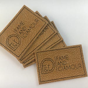 May include: A stack of rectangular, tan leather tags with the words "FAME AND GLAMOUR" printed in black. The tags have a textured surface and a black border. The logo includes the letters "F&G" inside a circle.