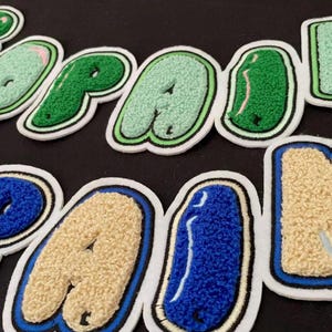Op de afbeelding: Een verzameling individuele getufte stoffen letters die de woorden COPAIN en PAIN vormen. De letters hebben een getextureerd, pluizig oppervlak met witte randen, uitgevoerd in tinten groen, blauw en crème in een vet, rond lettertype.