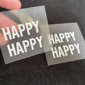 K&ouml;nnte beinhalten: Zwei transparente Plastikfolien mit dem Wort "HAPPY" in wei&szlig; darauf gedruckt. Die Folien liegen &uuml;bereinander.