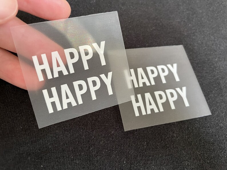K&ouml;nnte beinhalten: Zwei transparente Plastikfolien mit dem Wort "HAPPY" in wei&szlig; darauf gedruckt. Die Folien liegen &uuml;bereinander.