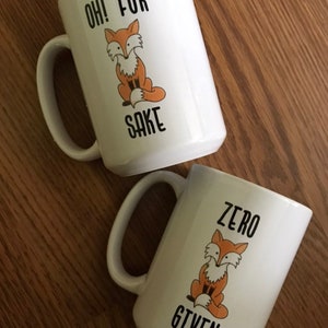 May include: Two white ceramic mugs with black text and a cartoon fox design. The first mug says "Oh! For Sake" and the second mug says "Zero Given".