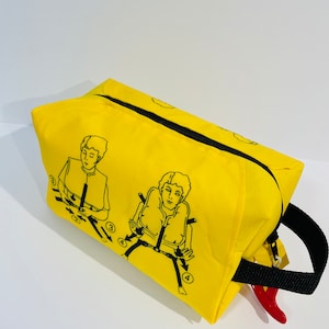 May include: A bright yellow rectangular bag with a black zip and handle. The bag has black line drawings illustrating life vest instructions. A red inflation device is visible. The bag is likely for safety equipment.
