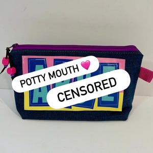 May include: A dark blue denim zippered pouch featuring a rectangular graphic patch with pink, yellow, and light blue block lettering. The zipper is purple with two pink beads on the pull. A pink fabric tab is attached to the right side of the pouch.