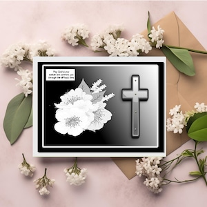 Op de afbeelding: Condoleancekaart met een zwart-wit bloemmotief en een zilveren kruis. De kaart bevat de tekst "May God's Love sustain and comfort you through this difficult time." Witte bloemen en een bruine envelop omringen de kaart.