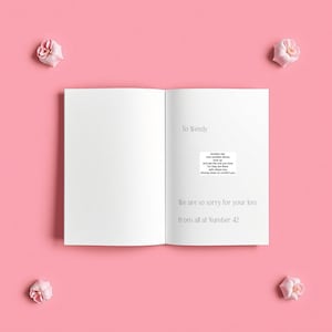 Op de afbeelding: Een open, witte kaart met de tekst "To Wendy" en "We are so sorry for your loss from all at Number 42." Er is ook een kleine, gedrukte boodschap opgenomen. Vier roze bloemen zijn rond de kaart gerangschikt op een roze achtergrond.