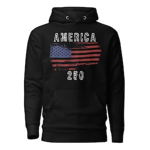 May include: Black colored hoodie featuring a America 250 with a distressed American Flag. The hoodie has a kangaroo pocket and a drawstring hood. The graphic is detailed with a distressed effect.