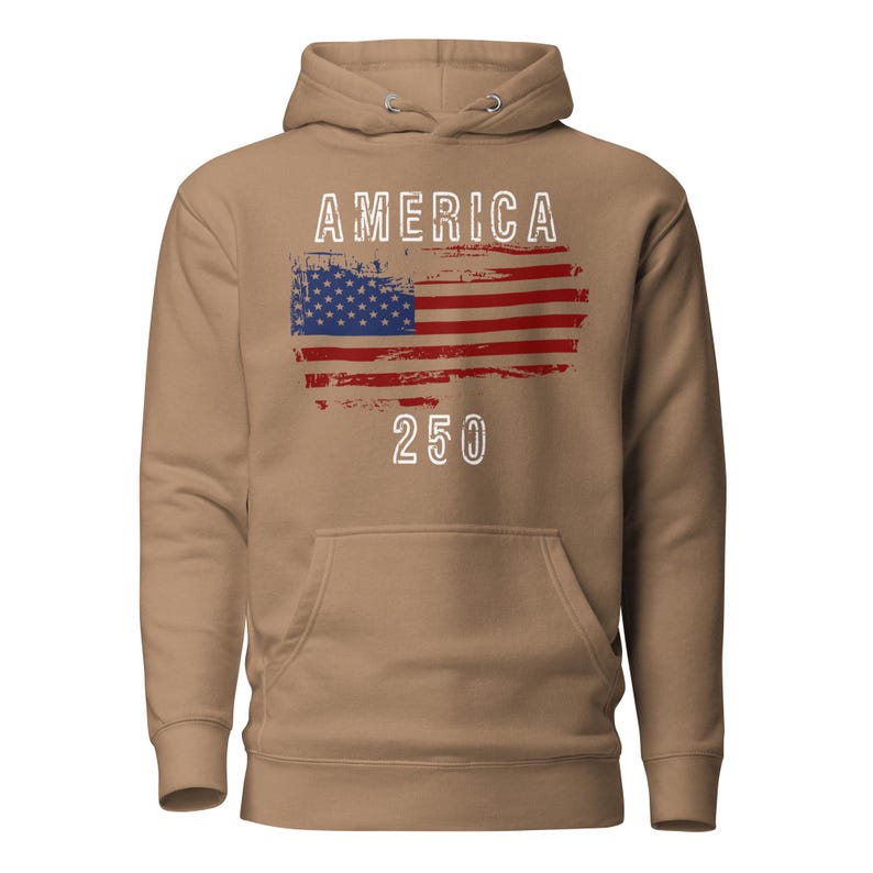May include: Latte colored hoodie featuring a America 250 with a distressed American Flag. The hoodie has a kangaroo pocket and a drawstring hood. The graphic is detailed with a distressed effect.