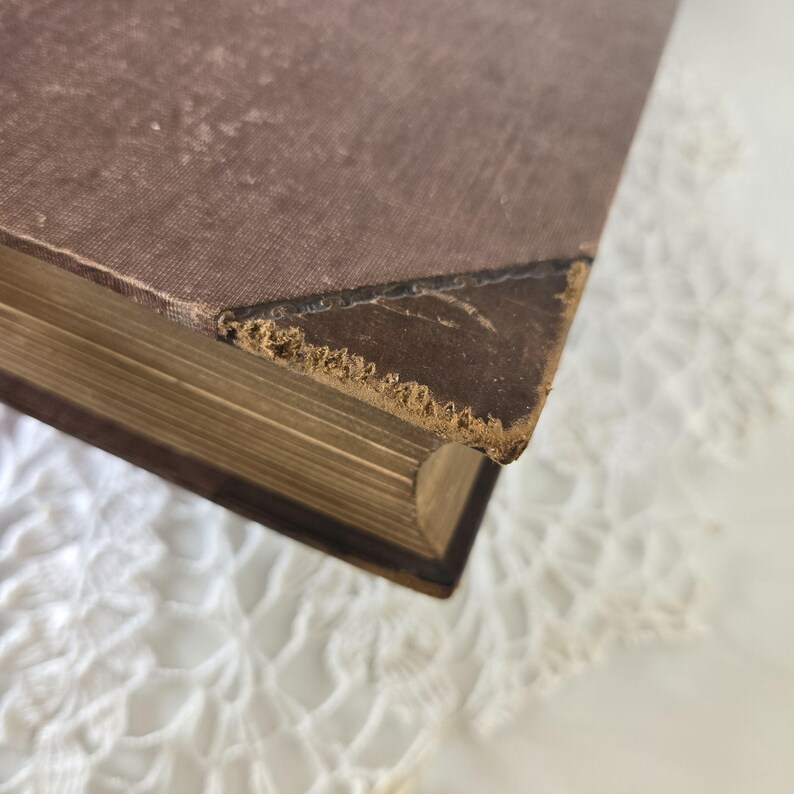 Op de afbeelding: Close-up van een vintage boek met een versleten, bruine kaft. De randen van de pagina's zijn verouderd en de hoeken vertonen slijtage. Het boek ligt op een wit, kantachtig oppervlak.