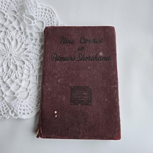May include: A maroon book with the title "New Course in Pitman's Shorthand" in black lettering. The book has a worn appearance, with a decorative emblem at the bottom. It rests on a white surface next to a white lace doily.