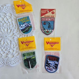 May include: Four iron-on patches featuring Canadian landmarks. The patches are on white cardstock with yellow borders and text that reads "A Product of Voyageur" and "Collection Mondial World Wide Collectors Series". The patches feature images of Banff, Jasper Sky Tram, Maligne Canyon, and Columbia Icefield.