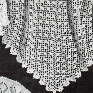 May include: A white, open-weave crochet shawl with a scalloped edge. The intricate pattern features a repeating geometric design. The shawl is displayed against a dark background, highlighting the delicate texture and craftsmanship.
