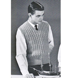 Puede incluir: Un chaleco de punto de cable gris claro con cuello en V, usado sobre una camisa blanca con cuello. El chaleco tiene un patrón texturizado. Una maqueta de barco está en primer plano.