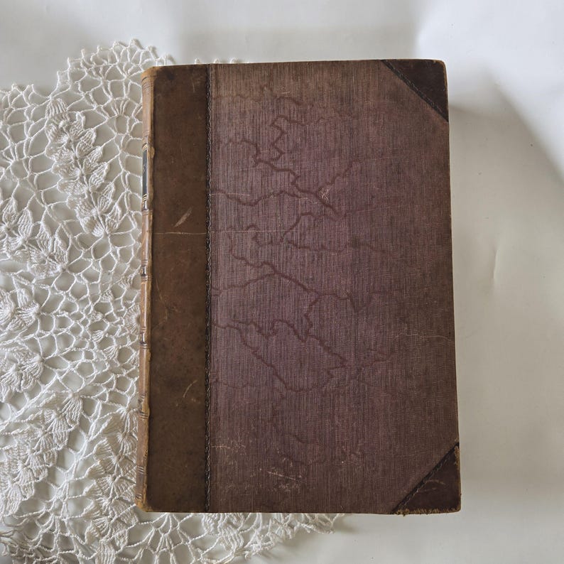 Op de afbeelding: Een vintage boek met een versleten, getextureerde kaft in bruin- en paars tinten. De rug is lichtbruin en de hoeken zijn verstevigd met donkerder bruin leer. De kaft heeft een uniek, gekraakt patroon.