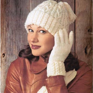 Könnte beinhalten: Ein cremefarbenes Strickmützen- und Handschuh-Set. Die Mütze hat ein Zopfmuster und eine Quaste. Die Handschuhe haben gerippte Bündchen. Die Frau im Bild trägt eine braune Lederjacke.