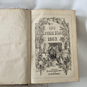Op de afbeelding: Open boek met de titel "The Leisure Hour 1863" in zwarte inkt. De omslag heeft een sierlijk bloemmotief en illustraties van mensen. De pagina's zijn verouderd met wat verkleuring, wat wijst op de vintage.