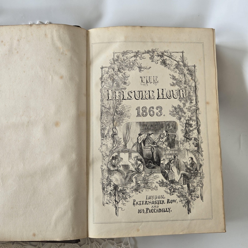 Op de afbeelding: Open boek met de titel "The Leisure Hour 1863" in zwarte inkt. De omslag heeft een sierlijk bloemmotief en illustraties van mensen. De pagina's zijn verouderd met wat verkleuring, wat wijst op de vintage.