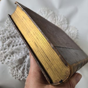 Op de afbeelding: Een vintage boek met een bruine leren kaft en goud vergulde randen. De pagina's van het boek zijn zichtbaar en de rug is licht versleten. Het boek wordt onder een hoek gehouden, waarbij de bladgoud details worden getoond.
