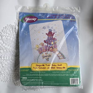 May include: A packaged Barney Sleepyville Train Baby Quilt kit. The quilt features an embroidered design of Barney and friends on a train. The package is blue with the Barney logo and text in English and French. Finished size is 34 x 43 inches.