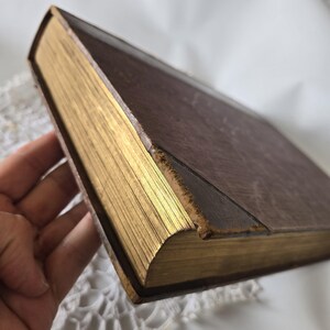 Op de afbeelding: Een vintage boek met een bruine leren kaft en goud vergulde randen. De pagina's van het boek zijn zichtbaar en de kaft vertoont tekenen van slijtage, wat duidt op de leeftijd. De gouden randen geven het boek elegantie.