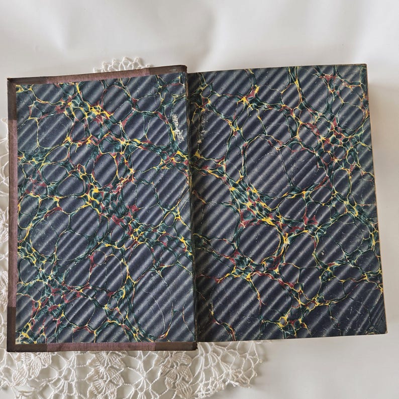 Op de afbeelding: Open antiek boek met gemarmerde papieren omslagen. De omslagen zijn donkerblauw met een diagonaal streepjespatroon en kleurrijke wervelingen van geel, rood en groen. De rug is donkerbruin.