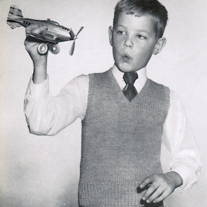 May include: A black and silver toy airplane with a propeller is held up by a person. The person is wearing a gray knitted vest over a white collared shirt and a dark tie. The image is in black and white.