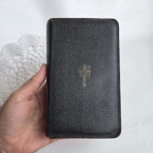 May include: A black book with a textured cover and a gold-coloured cross emblem. The book is rectangular and held in a hand. A white lace doily is visible in the background.