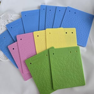 May include: A selection of Fiskars rubber texture plates in shades of blue, pink, yellow, and green. Each plate has a different embossed pattern, including lines, swirls, and floral motifs. The rectangular plates have a hole at the top for hanging.