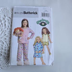 May include: A vintage Butterick sewing pattern, B5129, featuring a Cabbage Patch Kids design. The pattern shows illustrations of children's pajamas and a doll. The pattern's cover is white with pink and purple floral accents.