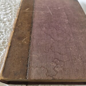 Op de afbeelding: Een vintage boek met een versleten, bruine leren rug en kaft. De kaft heeft een vervaagde, getextureerde kastanjebruine kleur en de rug heeft decoratieve metalen accenten. De randen van het boek vertonen tekenen van ouderdom en slijtage, wat suggereert dat het een antiek of een gekoesterd item is.