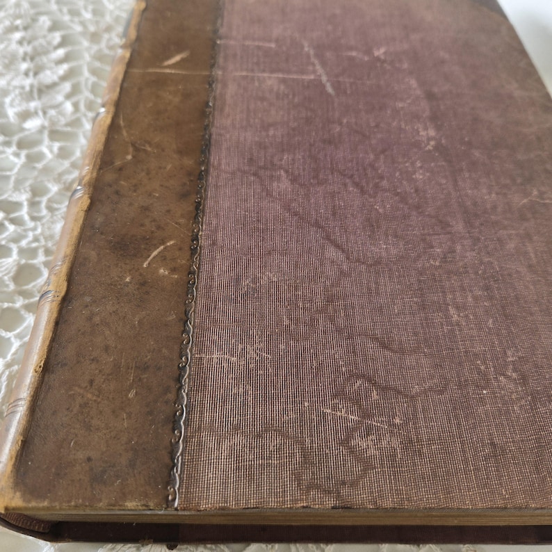 Op de afbeelding: Een vintage boek met een versleten, bruine leren rug en kaft. De kaft heeft een vervaagde, getextureerde kastanjebruine kleur en de rug heeft decoratieve metalen accenten. De randen van het boek vertonen tekenen van ouderdom en slijtage, wat suggereert dat het een antiek of een gekoesterd item is.