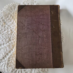 Op de afbeelding: Een vintage boek met een versleten, getextureerde kaft. De kaft is voornamelijk een gedempt paarsbruin met een gemarmerd patroon. De rug en hoeken zijn donkerbruin, wat een antieke uitstraling geeft. Het boek is waarschijnlijk een verzamelobject.