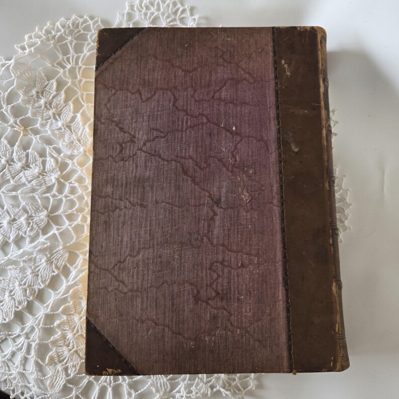 Op de afbeelding: Een vintage boek met een versleten, getextureerde kaft. De kaft is voornamelijk een gedempt paarsbruin met een gemarmerd patroon. De rug en hoeken zijn donkerbruin, wat een antieke uitstraling geeft. Het boek is waarschijnlijk een verzamelobject.