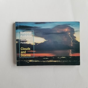 May include: A National Audubon Society Pocket Guide titled "Clouds and Storms." The cover features a dramatic image of a lightning storm over a city at dusk, with a vibrant orange and blue sky.