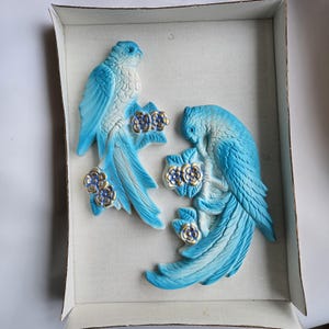 May include: Two light blue parrot wall decorations. Each parrot is perched on a branch with gold and silver flowers. The parrots are in a cardboard box.