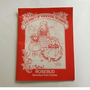 May include: A red booklet titled "A Garden of Smocking Designs" with illustrations of three dolls in smocked dresses. The booklet is labeled "ROSEBUD Smocked Doll Clothes".