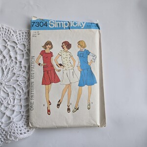 Peut inclure: Patron de couture Simplicity vintage, taille 12, avec des illustrations de trois modèles de robes différents. L'enveloppe du patron est blanc cassé avec du texte et des illustrations bleus. Le texte "7304 Simplicity" est visible en haut.
