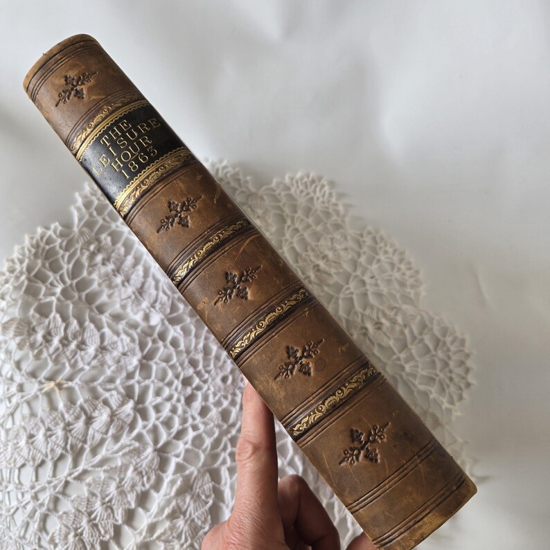 Op de afbeelding: Een vintage, bruin ledergebonden boek met de titel "The Leisure Hour 1863". Het boek heeft reli&euml;f bloemmotieven en gouden accenten. De rug is donker met gouden letters. Het boek wordt door een hand vastgehouden.