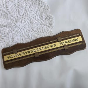 May include: A vintage wooden scoring board with brass detailing. The board has two parallel metal rods and two brass plates with numbers. The numbers are printed in a classic font, ranging from 0 to 100. The board is set against a white lace background.