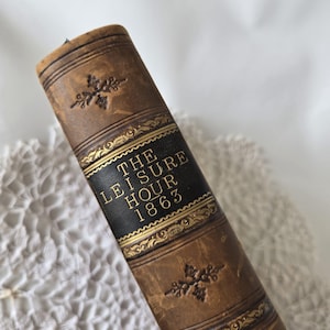 Op de afbeelding: Close-up van een vintage boekrug. Het boek is bruin met gouden accenten en de titel "THE LEISURE HOUR 1863" in gouden letters op een zwart paneel. Decoratieve bloempatronen zijn in reli&euml;f op de rug.