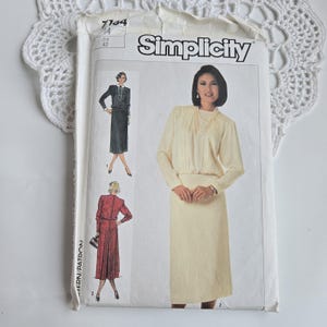 May include: A vintage Simplicity sewing pattern featuring illustrations of women's dresses. The pattern envelope is white with the Simplicity logo and text. The pattern number is 7764, with a size 14 and Eur 42. The image shows a cream-colored dress.