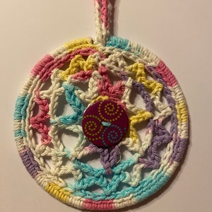 May include: A crocheted circle ornament with a pink, yellow, blue, and white color scheme. The ornament has a button in the center with a purple background and a yellow and blue swirl design.