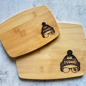 May include: Two wooden cutting boards with a funny saying and a cartoon image of a person wearing a hat and glasses. The text on the boards says "Oh Fudge!"