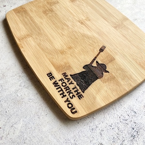 May include: A brown wooden cutting board with the text "May the forks be with you" and a silhouette of a figure holding a fork.