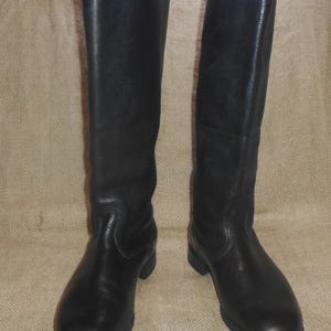 May include: A pair of black leather knee-high boots. The boots have a smooth, polished appearance and a rounded toe. They feature a low heel and are displayed upright, highlighting their full length and design. The boots are made of leather.